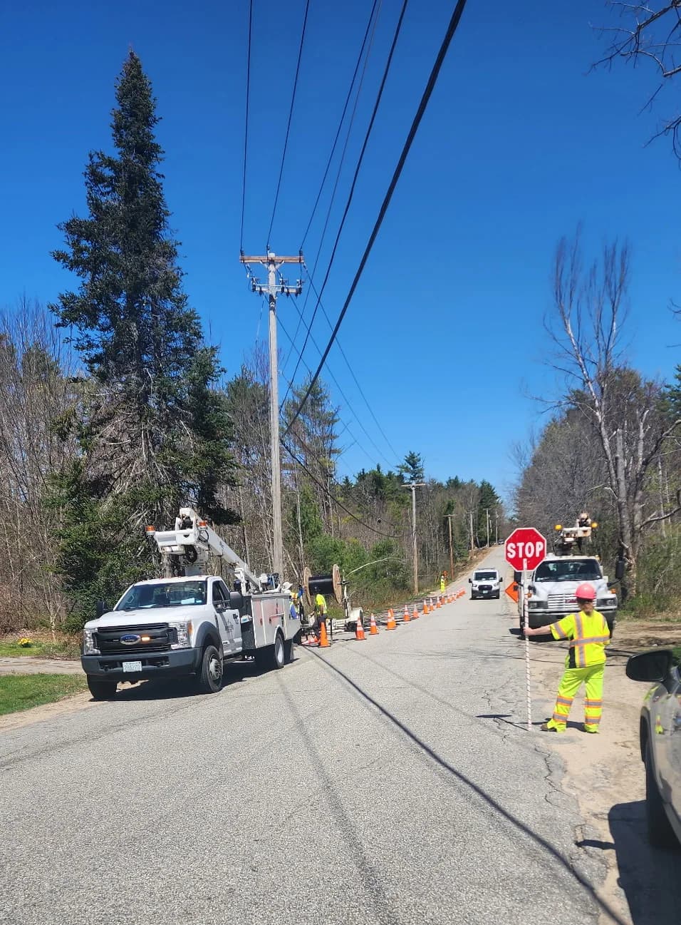 A certified flagger holding a STOP paddle at a cone taper while aerial crews work the line behind