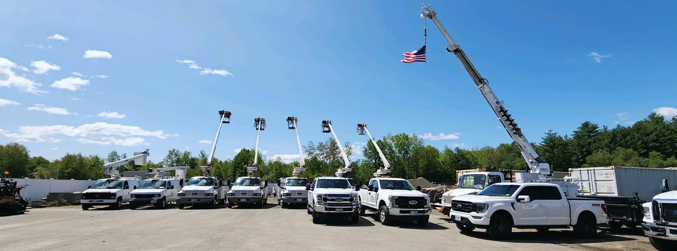 Six bucket trucks and pickups lined up in the MBG yard, an American flag flying from the boom of the largest