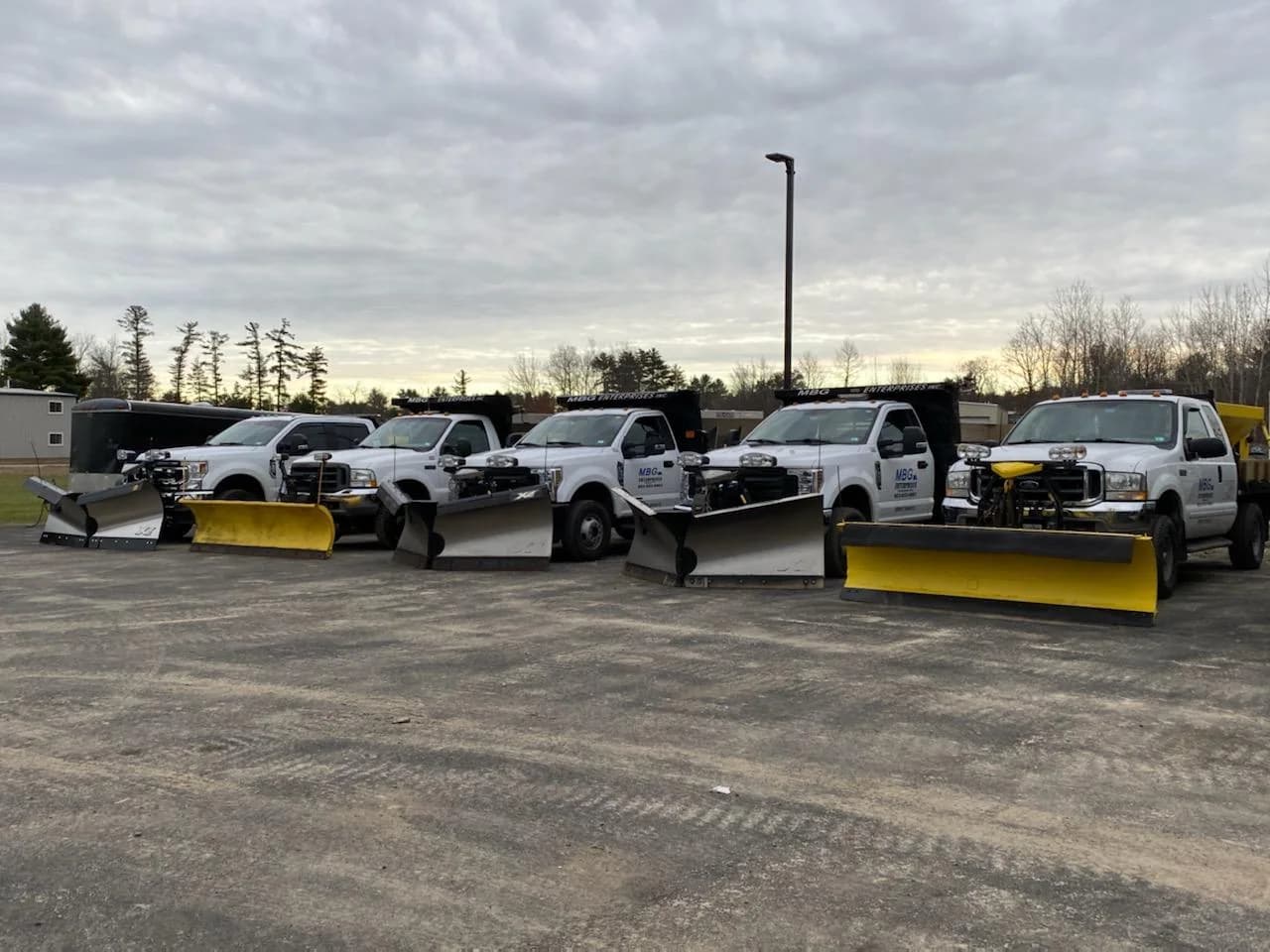 Five MBG plow trucks lined up at dusk ahead of a storm