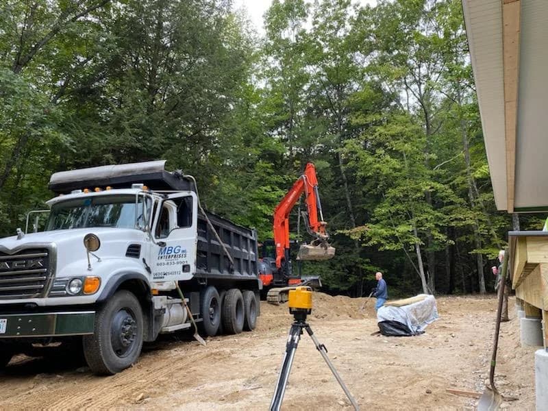 An MBG dump truck and compact excavator setting grade beside a house under construction, laser level in the foreground