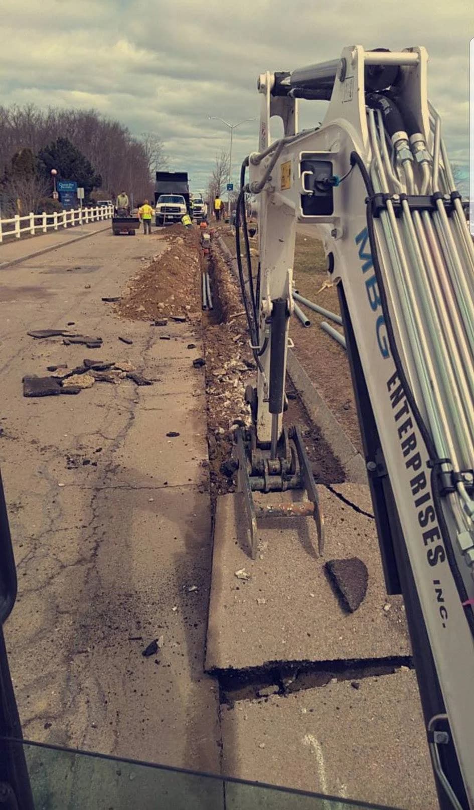 An MBG mini excavator cutting a conduit trench along a roadway shoulder, crew and dump truck ahead