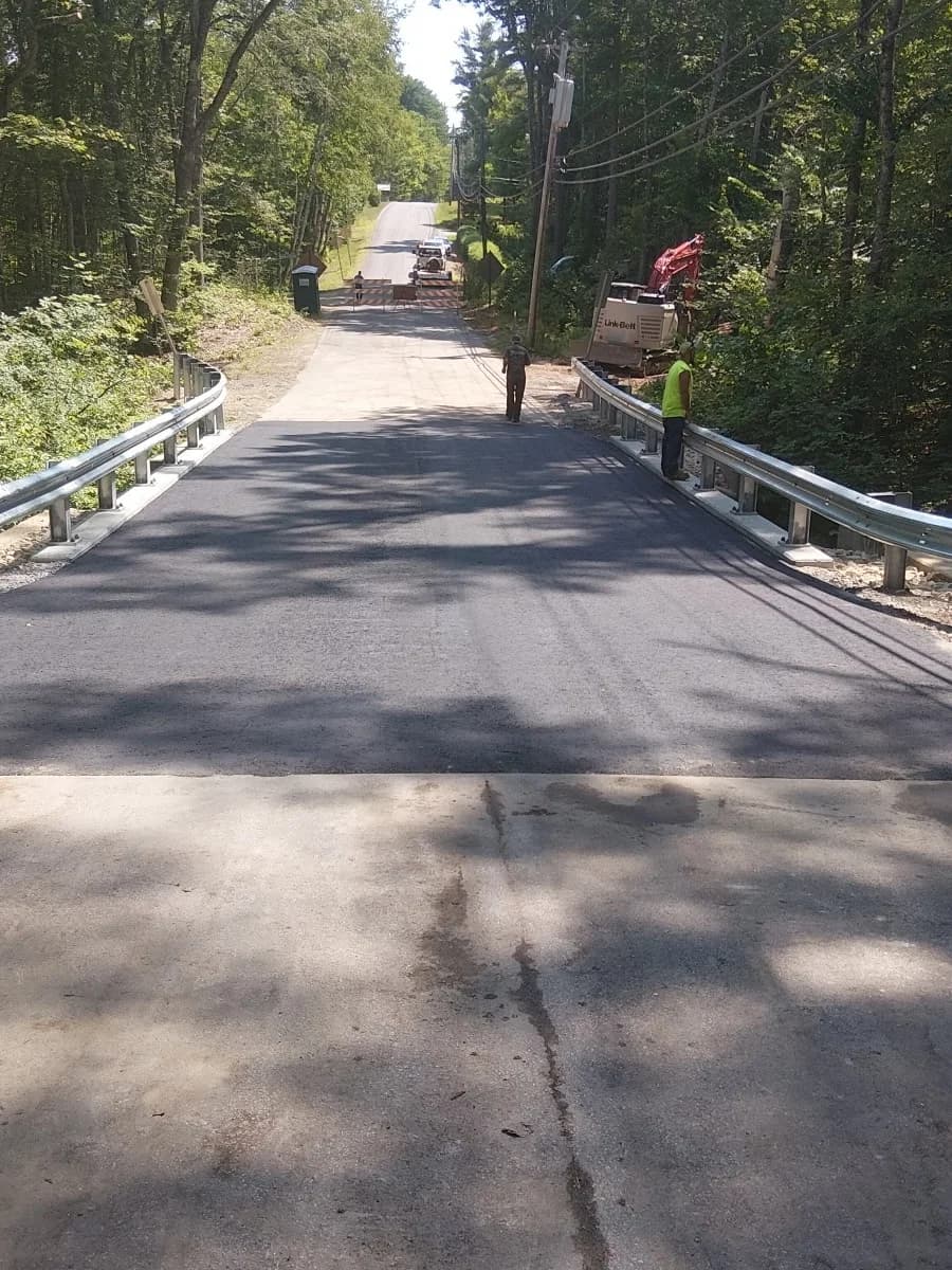 A freshly repaved town road between guardrails, crew and excavator still on site